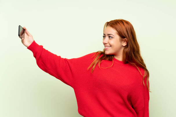携帯電話で自分撮りを作る孤立した緑の背景の上にセーターを持つティーンエイジャー赤毛の女の子 - 写真・画像