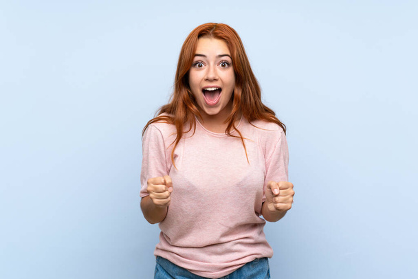 Teenager redhead girl over isolated blue background celebrating a victory in winner position - 写真・画像