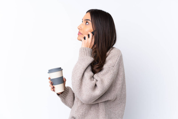 Νεαρή μελαχρινή γυναίκα πάνω από απομονωμένο λευκό φόντο κρατώντας καφέ για να πάρει μακριά και ένα κινητό - Φωτογραφία, εικόνα