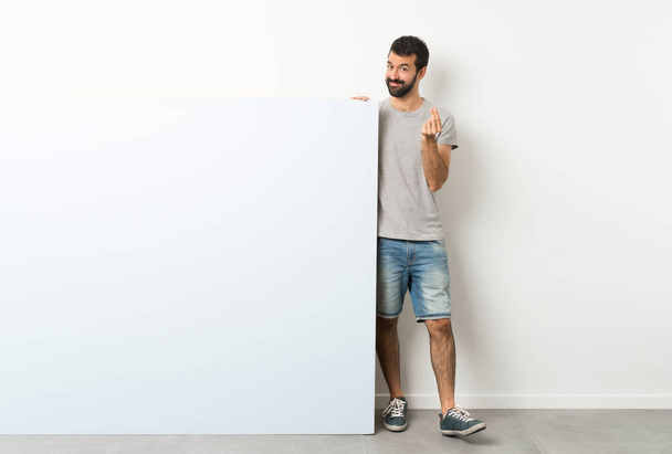 Young handsome man with beard holding a big blue empty placard making money gesture - 写真・画像