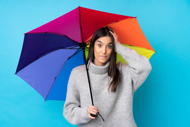 Young brunette woman holding an umbrella over isolated blue wall with surprise facial expression - 写真・画像