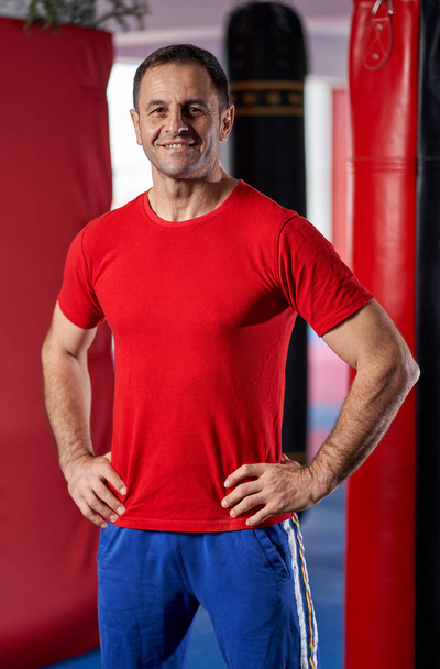 Kickboxing coach posing in his gym with punchbags in background - Foto, imagen