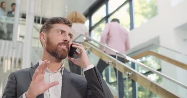 Close up of handsome businessman talking on his smartphone and standing in office building. Male office worker in suit having phone conversation. Concept of business and communication. - Video