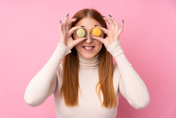 Joven pelirroja sobre fondo rosa aislado usando coloridos macarrones franceses como gafas
 - Foto, Imagen