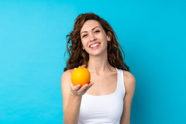 Молодая женщина с вьющимися волосами держит апельсин на изолированном синем фоне
 - Фото, изображение