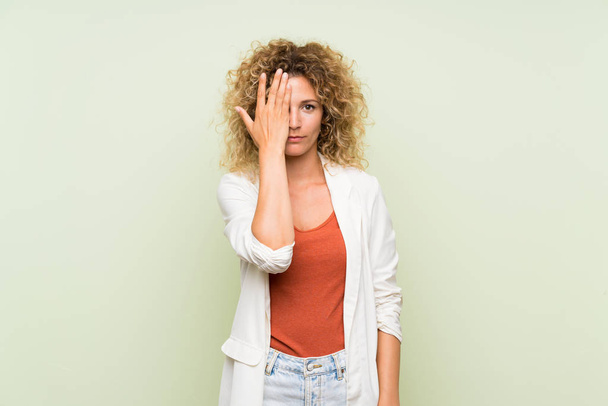 Mladá blondýna žena s kudrnaté vlasy přes izolované zelené pozadí pokrývající oko rukou - Fotografie, Obrázek