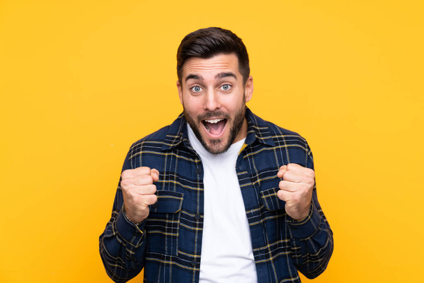 Young handsome man with beard over isolated yellow background celebrating a victory in winner position - Φωτογραφία, εικόνα