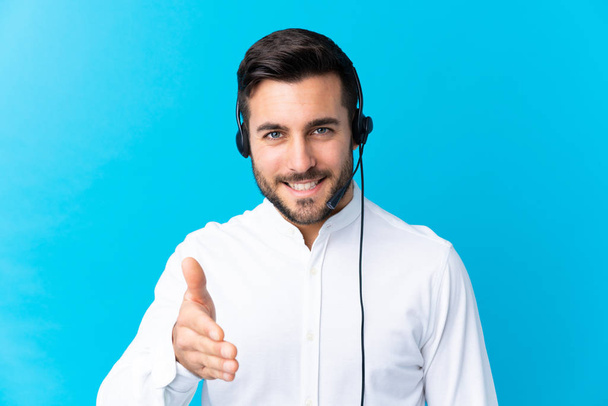Telemarketer man werken met een headset over geïsoleerde blauwe achtergrond handdrukken na een goede deal - Foto, afbeelding