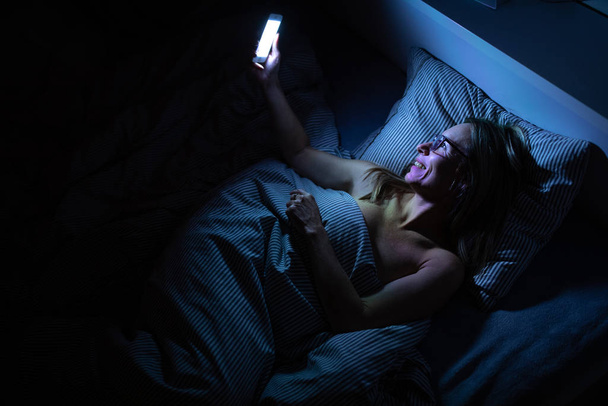 Pretty, middle-aged woman using her cell phone in bed  - Fotografie, Obrázek