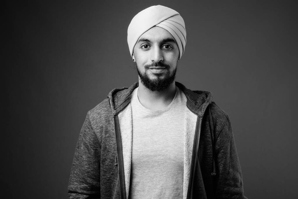 Studio shot del giovane Sikh indiano barbuto che indossa giacca su sfondo grigio in bianco e nero - Foto, immagini