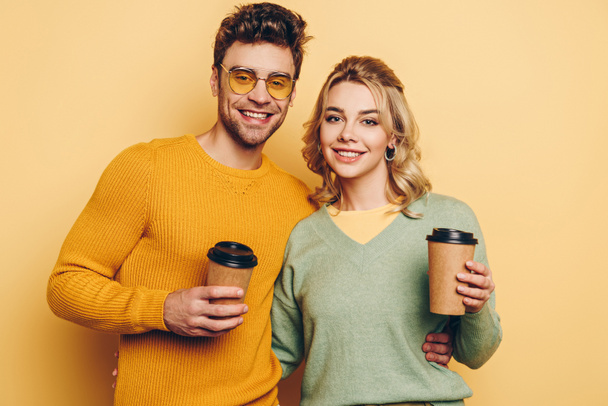 cheerful couple looking at camera while holding coffee to go on yellow background - Fotografie, Obrázek