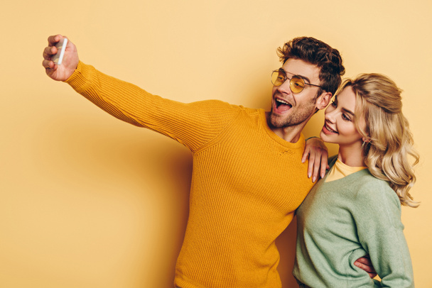 cheerful man taking selfie on smartphone with pretty girlfriend on yellow background - Photo, Image