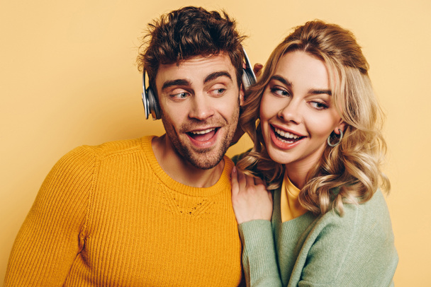 happy girl embracing boyfriend while listening music in wireless headphones together on yellow background - Zdjęcie, obraz
