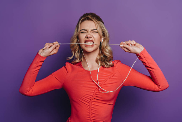 thrilled girl biting wire of earphones while listening music on purple background  - Fotó, kép