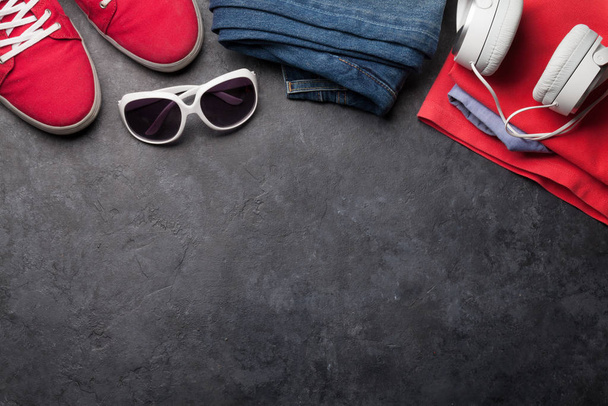 Kleding en accessoires. Sneakers, jeans en koptelefoon. Stedelijke outfit voor alledaagse of reis vakantie op stenen achtergrond met kopieerruimte. Bovenaanzicht vlak lay - Foto, afbeelding