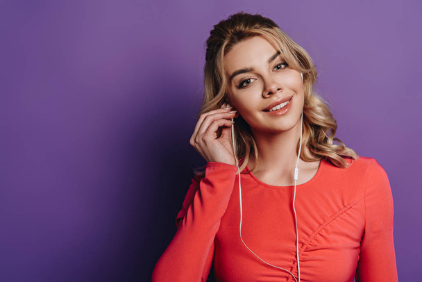 smiling girl listening music in earphones and looking at camera on purple background - Foto, Imagem