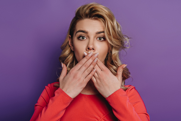 shocked girl covering mouth with hands while looking at camera on purple background - Foto, afbeelding