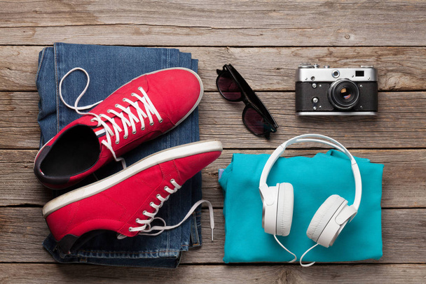 Oblečení a doplňky. Tenisky, džíny, foťák a sluchátka. Městské oblečení pro každodenní nebo cestování dovolenou na dřevěném pozadí. Horní pohled rovný - Fotografie, Obrázek