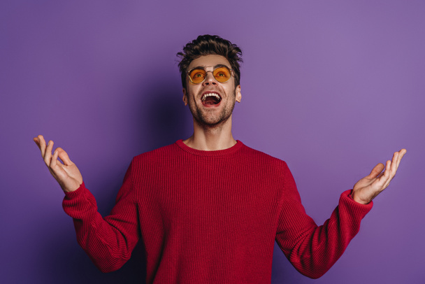 happy man looking up while standing with open arms on purple background - Photo, Image