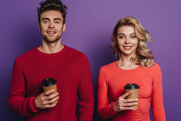 sonriente pareja mirando a la cámara mientras sostiene el café para ir sobre fondo púrpura
 - Foto, Imagen