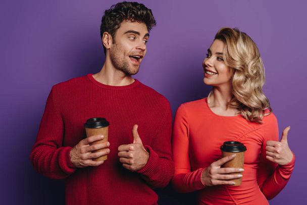 feliz pareja mostrando los pulgares hacia arriba mientras sostiene el café para ir sobre fondo púrpura
 - Foto, Imagen