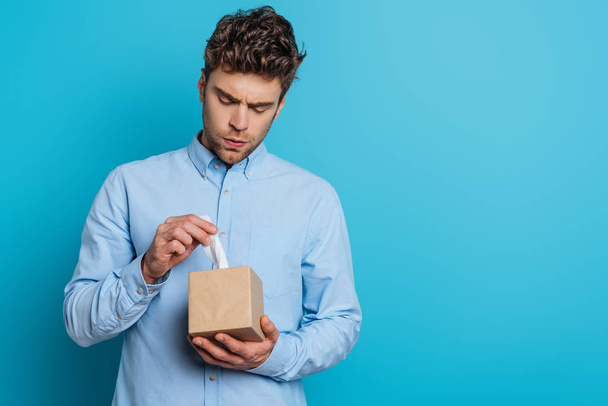 upset young man taking paper napkin from pack on blue background - Φωτογραφία, εικόνα