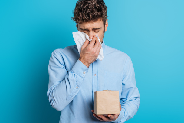 diseased young man sneezing in paper napkin on blue background - Foto, Imagem