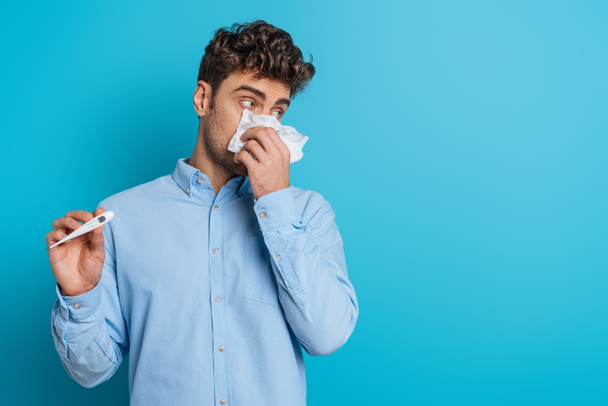 joven enfermo limpiando la nariz con servilleta de papel y mostrando termómetro sobre fondo azul
 - Foto, Imagen