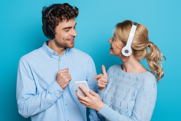 positive couple in headphones looking at each other while girl pointing with finger at smartphone on blue background - Fotó, kép