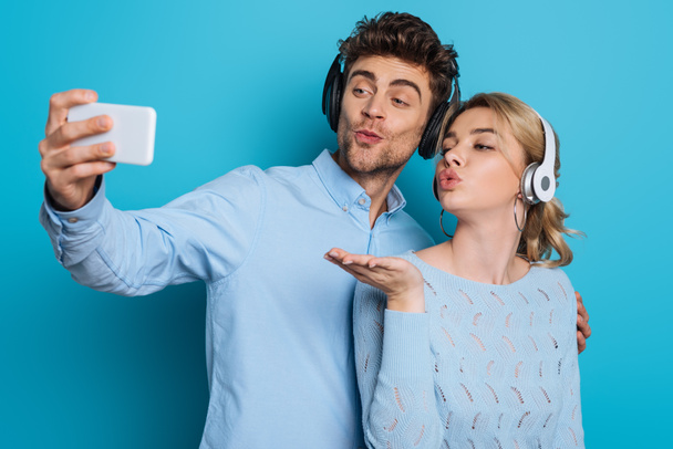 Junge Männer und Frauen mit drahtlosen Kopfhörern senden Luftküsse, während sie ein Selfie auf dem Smartphone auf blauem Hintergrund machen - Foto, Bild