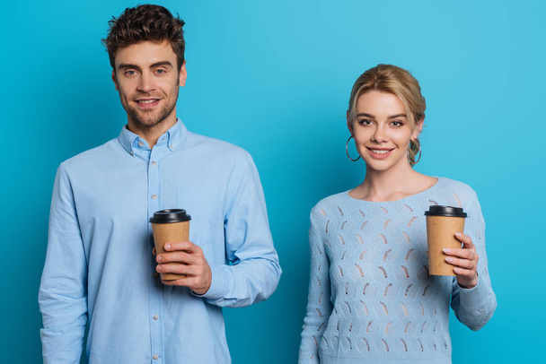 positive man and woman holding disposable cups while smiling at camera on blue background - Photo, Image