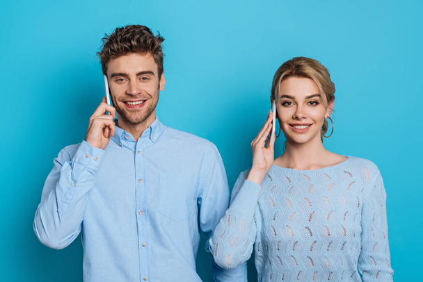 young man and woman smiling at camera while talking on smartphones on blue background - Photo, image