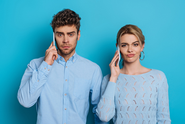 skeptical man and woman looking at camera while talking on smartphones on blue background - Фото, зображення