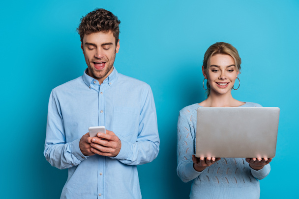 cheerful man chatting on smartphone near girlfriend holding laptop on blue background - Photo, Image