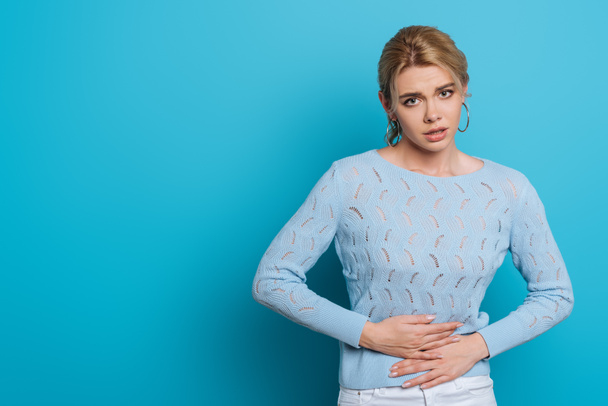 upset girl touching stomach while suffering from abdominal pain on blue background - Fotó, kép