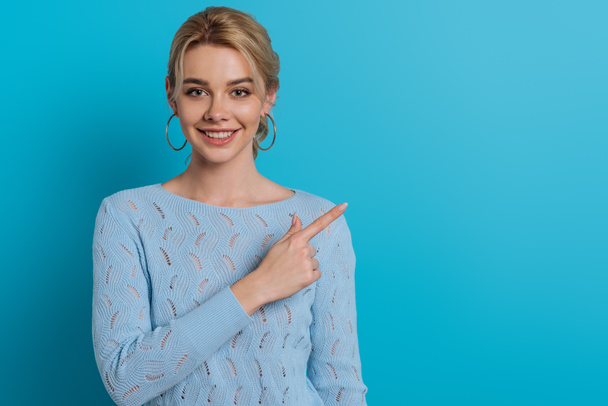 pretty girl smiling at camera while pointing with finger on blue background - Foto, immagini