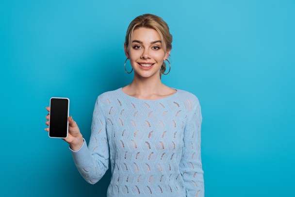 cheerful girl smiling at camera while showing smartphone with blank screen on blue background - Фото, изображение
