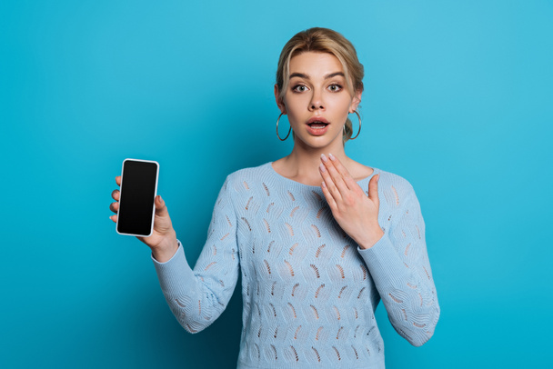 wstrząśnięta dziewczyna trzymając rękę blisko twarzy podczas pokazywania smartfona z pustym ekranem na niebieskim tle - Zdjęcie, obraz