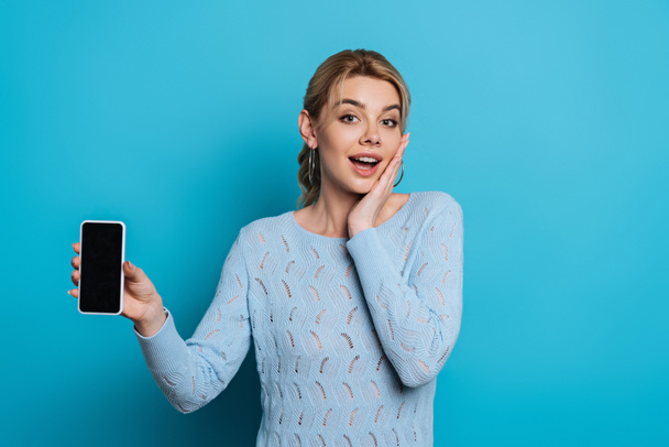 überraschtes Mädchen berührt Gesicht, während Smartphone mit leerem Bildschirm auf blauem Hintergrund zeigt - Foto, Bild