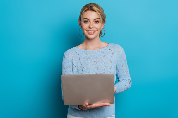 happy girl smiling at camera while using laptop on blue background - Photo, Image