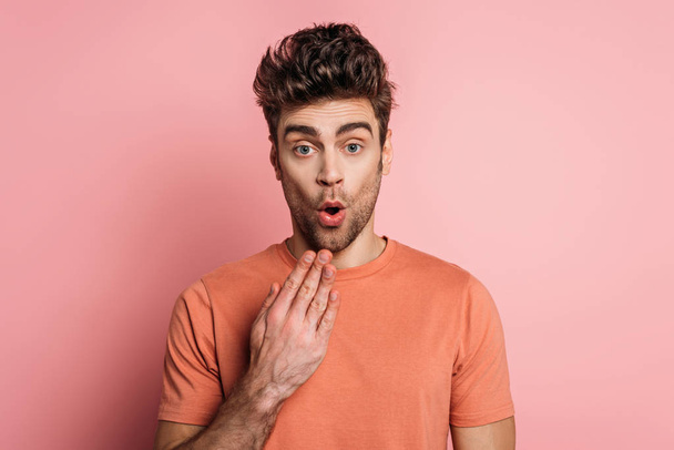 shocked man holding hand near mouth while looking at camera on pink background - Zdjęcie, obraz