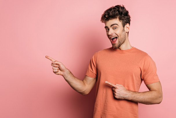 amazed young man pointing with fingers while looking at camera on pink background - Fotografie, Obrázek
