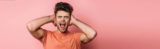 panoramic shot of irritated young man shouting and covering ears with hands on pink background - Fotoğraf, Görsel