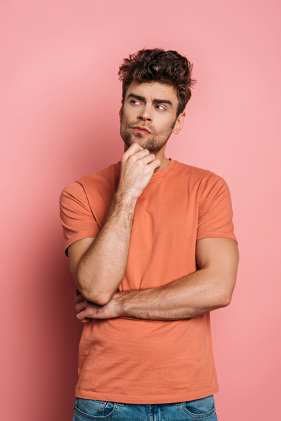 thoughtful young man touching chin while looking away on pink background - Фото, изображение