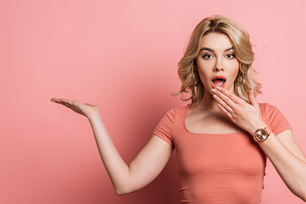 shocked girl covering mouth with hand while pointing with finger on pink background - Valokuva, kuva
