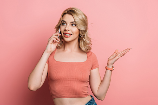 positive girl standing with open arm while talking on smartphone on pink background - Foto, Imagen