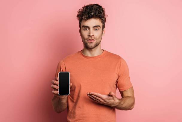serious young man pointing with hand at smartphone with blank screen on pink background - Foto, Imagem