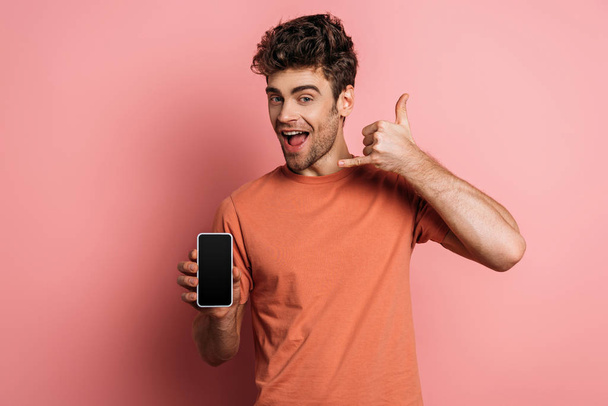 vidám fiatalember bemutató lehetővé teszi, hogy inni gesztus, miközben gazdaság smartphone üres képernyő rózsaszín háttér - Fotó, kép