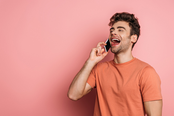 alegre joven riendo mientras habla en el teléfono inteligente sobre fondo rosa
 - Foto, Imagen