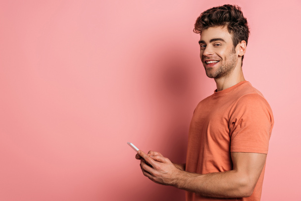 fröhlicher junger Mann lächelt in die Kamera, während er auf rosa Hintergrund mit dem Smartphone chattet - Foto, Bild